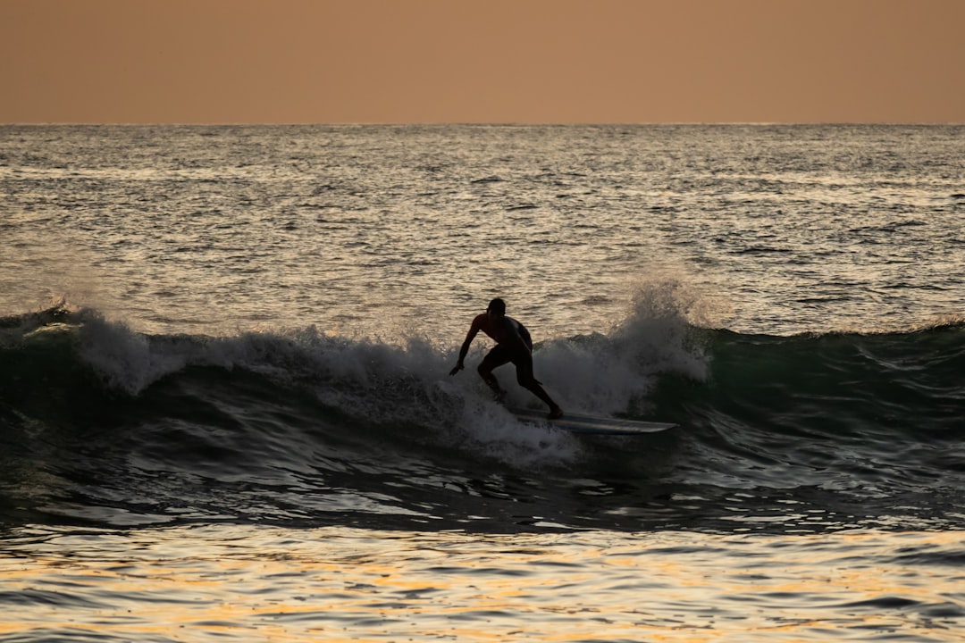 Sunset Surfing in Nosara - This stunning photograph captures the exhilarating thrill of surfing in the warm waters of Nosara, Costa Rica. The silhouette of the surfer is set against the golden hues of a magnificent sunset, as he rides the waves with incredible skill and grace. The natural beauty of the coastline, combined with the excitement of the sport, makes for an unforgettable experience. This photograph perfectly captures the sense of adventure and joy that surfing in Nosara offers. by Fitness and Nature a man riding a wave on top of a surfboard